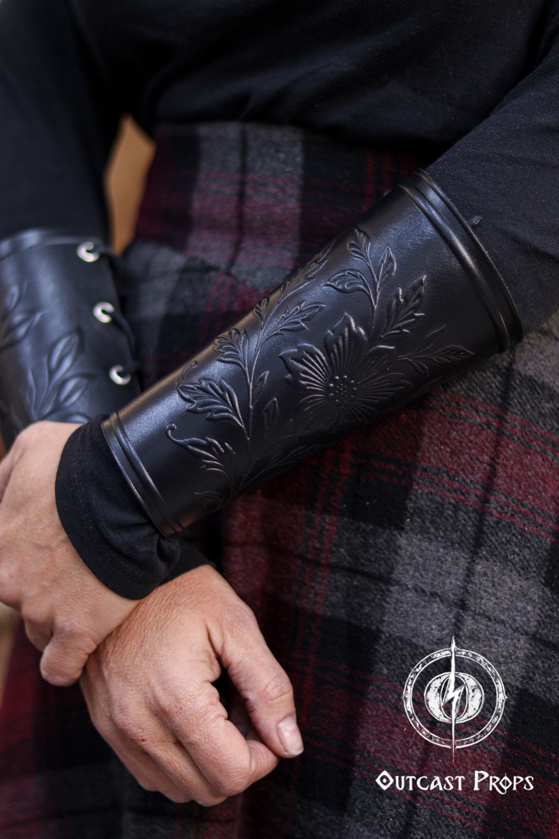 The Wildflower leather bracer in black is shown worn on the forearm with hands gently crossed, displaying the embossed floral design against a plaid skirt. This renaissance faire accessory is ideal for larp cosplay accessory outfits, elven herbalist witch characters and fantasy costumes seeking elegant botanical leather details. The dark tone emphasizes the carved pattern while maintaining a refined handcrafted appearance.