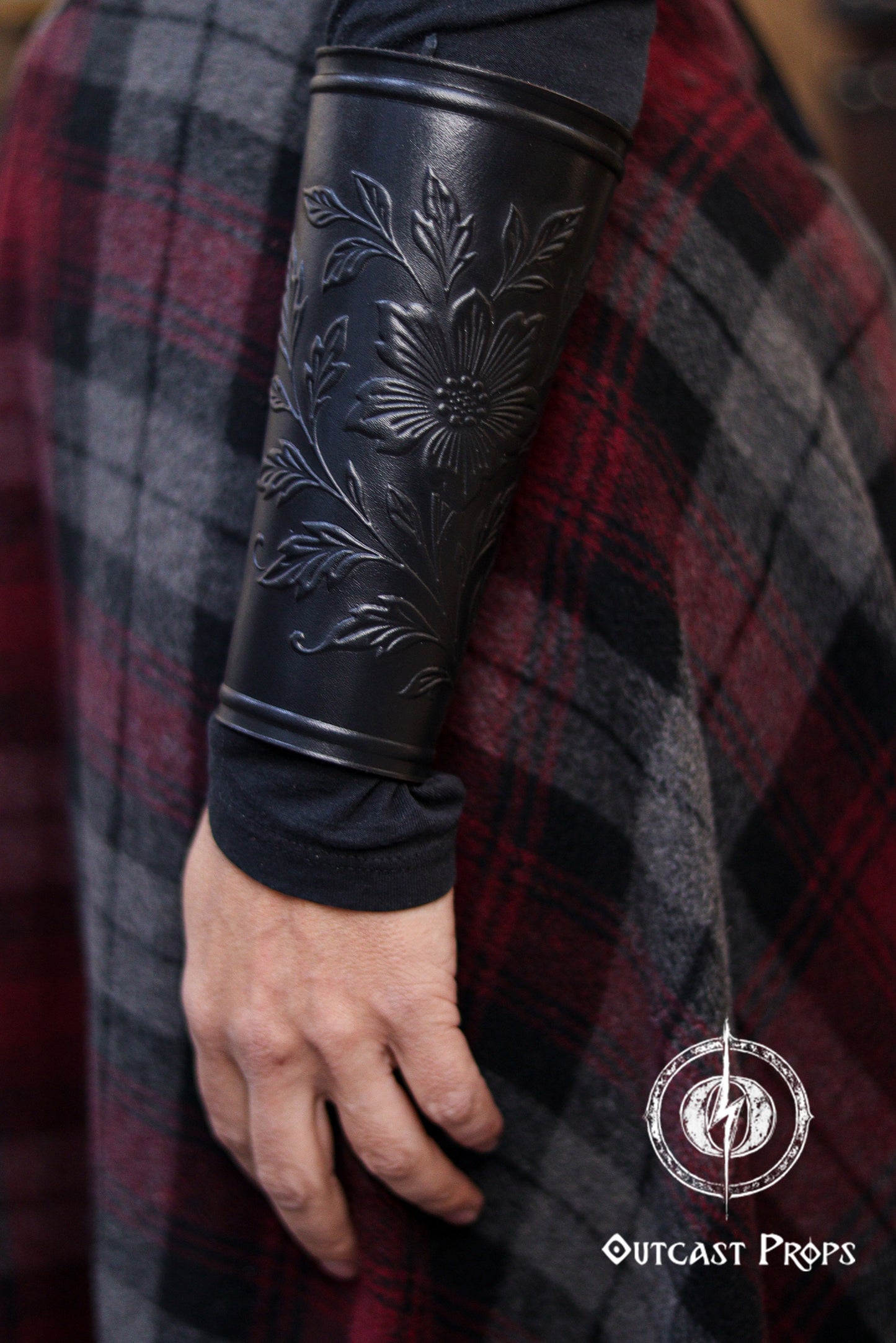 A side view of the Wildflower leather bracer in black shows the floral embossed artwork along the curved forearm shape. The bracer contrasts with the plaid skirt and dark top, creating a classic renaissance faire accessory look. Suitable for larp cosplay accessory costumes, elven herbalist witch roles and fantasy characters who want natural motifs in handcrafted leather gear. The image highlights texture, shape and fit.