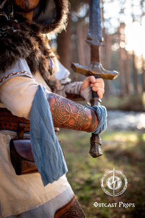 Close-up of a warrior gripping a sword with both hands, showcasing brown leather bracers with tree of life embossing. The outfit combines linen, fur, and blue wraps, ideal for larp barbarian gear, viking costume armor, leather vambrace handmade, or renaissance cosplay outfit. Forest lighting highlights the texture and aged finish of the embossed leatherwork. Yggdrasil Viking leather bracer world tree vambrace for larps cosplay yule high-quality full-grain vegetable tanned leather embossed armor