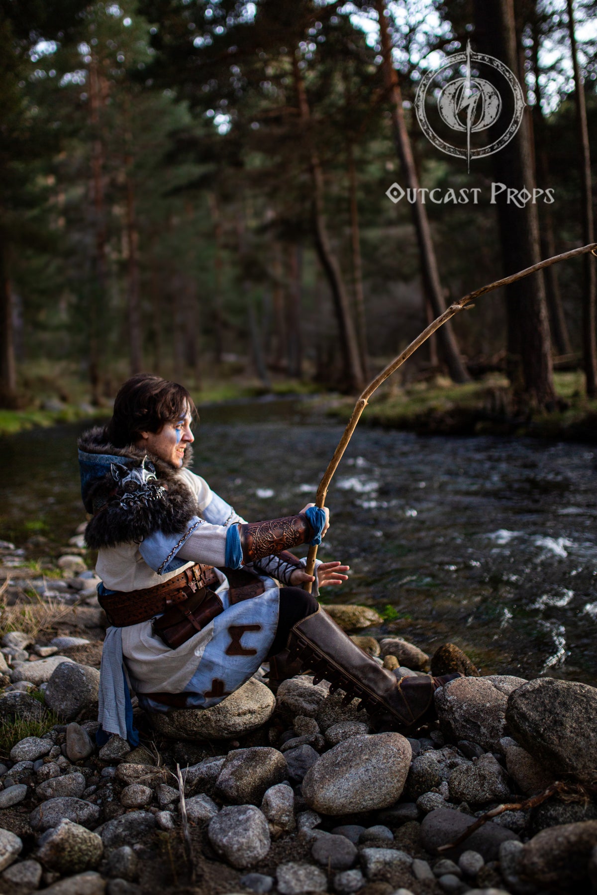 A fantasy warrior in fur cloak and linen robes fishes beside a forest stream, using a wooden rod. He wears brown leather bracers with Norse engraving, ideal for barbarian cosplay, larp ranger armor, and nordic costume accessories. The soft sunset light enhances the hand-embossed detail on the bracers, suggesting a moment of calm before action in a fantasy LARP scenario.
