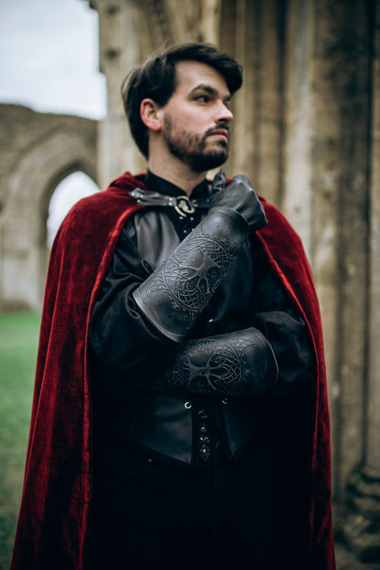 A fantasy LARP warrior wearing dark leather clothing and a red velvet cloak shows off black Yggdrasil leather bracers embossed with Norse world tree details. These long bracers, perfect for viking costume, leather armor, or medieval cosplay, offer both protection and character depth. Ideal for renaissance fairs, Geralt cosplay, or as female leather armor accessories. Model stands in front of a ruined stone archway, crossing arms with solem