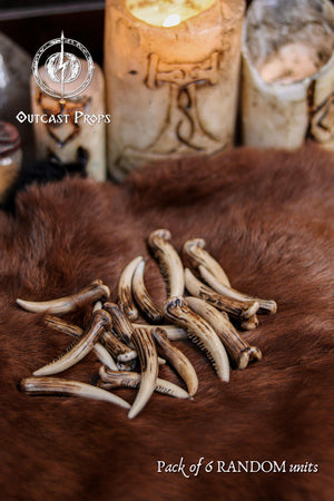 A pile of resin fang tusks displayed on brown fur with carved candles and fantasy props in the background, creating an atmospheric scene. The tusks feature aged bone tones and unique shapes ideal for cosplay prop supplies, diy fantasy crafting, tribal accessories, shaman costumes, creature design elements and LARP props. Random pack of six tusks measuring 6 to 7 cm, hand painted with natural variation.