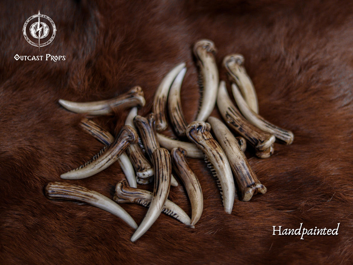 A scattered collection of hand painted resin fang tusks laid on brown fur, showing aged bone coloration and natural variations. These tusks are ideal for cosplay prop supplies, tribal accessories, shaman or druid costume building, diy fantasy crafting, creature design, rustic talismans, and handcrafted decorations. Each piece is 6 to 7 cm long, part of a random mix of shapes and textures.