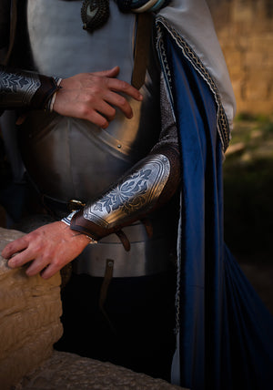 Close-up of a medieval knight costume showing detailed engraved leather bracers with silver metal plates, floral motifs, and leather straps. Perfect for fantasy cosplay, LARP armor, or noble character outfits. The texture of the armor and natural light enhance the historical look. Ideal accessory for renaissance fair or fantasy roleplay events.