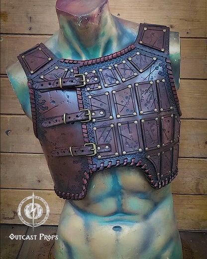 A handmade leather studded armor displayed on a stand, featuring chest plates with studs and an adjustable buckle system,. The armor has a brown distressed finish and is positioned against a backdrop with autumn leaves. A handmade leather studded armor displayed on a wooden stand with a fur cape, featuring brown leather plates with studs, and a corded edge finish. Barbarian studded leather chest armor with rabbit fur cape attachment shown on mannequin. Perfect for fantasy LARP, Viking
