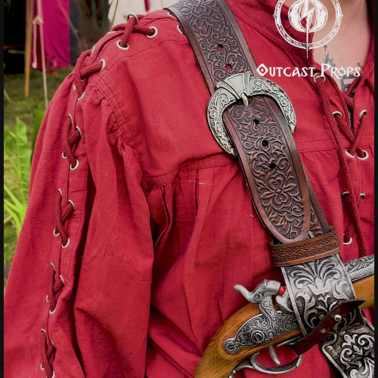 Pirate baldric for 4 flintlock pistols and a sword, worn crossbody over a red shirt. Inspired by Blackbeard and Anne Bonny, this silver-engraved leather shoulder belt is perfect for Jack Sparrow costume, Black Sails fans, LARP, renaissance fair, cosplay or Pirate Weekend. Handmade with vegetable-tanned leather. Premium pirate accessory for reenactment and fantasy events, ideal for themed photoshoots. Man in a red outfit with props, standing outdoors with greenery and tents in the background.