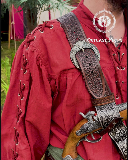 Pirate baldric for 4 flintlock pistols and a sword, worn crossbody over a red shirt. Inspired by Blackbeard and Anne Bonny, this silver-engraved leather shoulder belt is perfect for Jack Sparrow costume, Black Sails fans, LARP, renaissance fair, cosplay or Pirate Weekend. Handmade with vegetable-tanned leather. Premium pirate accessory for reenactment and fantasy events, ideal for themed photoshoots. Man in a red outfit with props, standing outdoors with greenery and tents in the background.