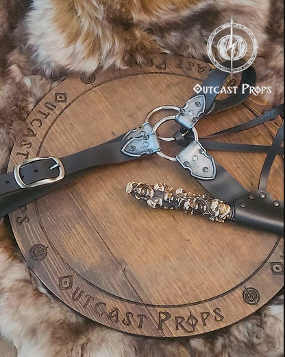 A black and silver leather wand holster with dual metal rings and riveted construction. Styled for Voldemort-inspired cosplay or LARP magic duels. Crafted for comfort and realism in wizard costumes, especially for Harry Potter fans recreating Death Eater aesthetics.Magic-style leather belt with decorative elements on a wooden surface, featuring Outcast Props branding.