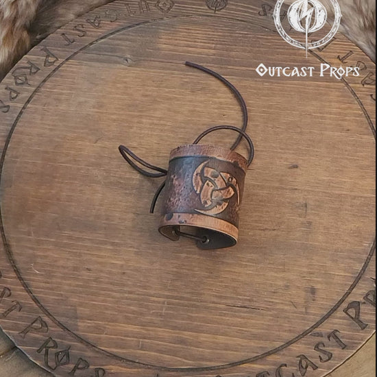 Leather cuff bracelet with the Three Horns of Odin design, placed against a backdrop with candles and decorative foliage. Horns of Odin Leather Cuff Bracelet, Viking Bracer, leather embossed wristband, stamped viking pattern for LARP, cosplay and Fantasy fairs