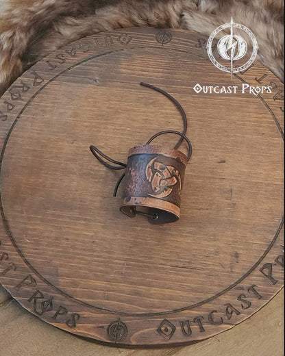 Leather cuff bracelet with the Three Horns of Odin design, placed against a backdrop with candles and decorative foliage. Horns of Odin Leather Cuff Bracelet, Viking Bracer, leather embossed wristband, stamped viking pattern for LARP, cosplay and Fantasy fairs