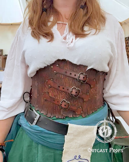 Person in a fantasy costume with 'Outcast Props' logo in the corner. Person wearing a detailed leather corset with decorative elements, branded 'Outcast Props'. Underbust Wood Leather Corset. Herbalist corset. Druid corset. Elven Corset. Ivy Corset.