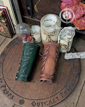 Two leaf elven bracers displayed on a carved wooden board with the Outcast Props logo. Their embossed leaf pattern and aged leather finish show craftsmanship ideal for renaissance faire costumes, larp characters, elves, witches, herbalists or fae themed outfits. These leather bracers also complement roman laurel or Caesar cosplay thanks to the leaf motif. Laced fastening and warm tones highlight their elegant look.