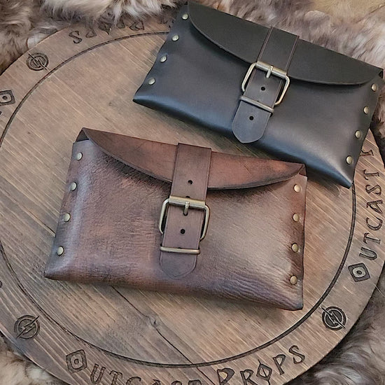 Medieval leather pouch with buckle closure placed on rustic wooden surface. Handmade Renaissance fair leather bag with natural brown tone, brass studs and curved flap. Perfect for LARP costume accessory or fantasy belt bag. Shows artisan texture of vegetable-tanned leather. Ideal for viewers needing a detailed tactile and visual description.