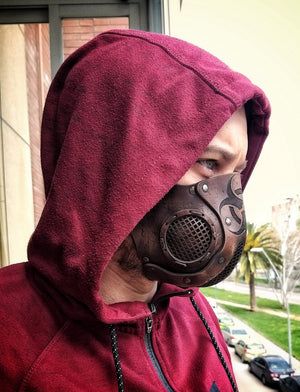 A person wearing a leather face mask with a hood, featuring a biohazard symbol and adjustable straps, in front of a background of foliage. Leather Face Mask with filter pocket Biohazard sign Post Apocalyptic mask for use with disposable face masks Paper towel or filter