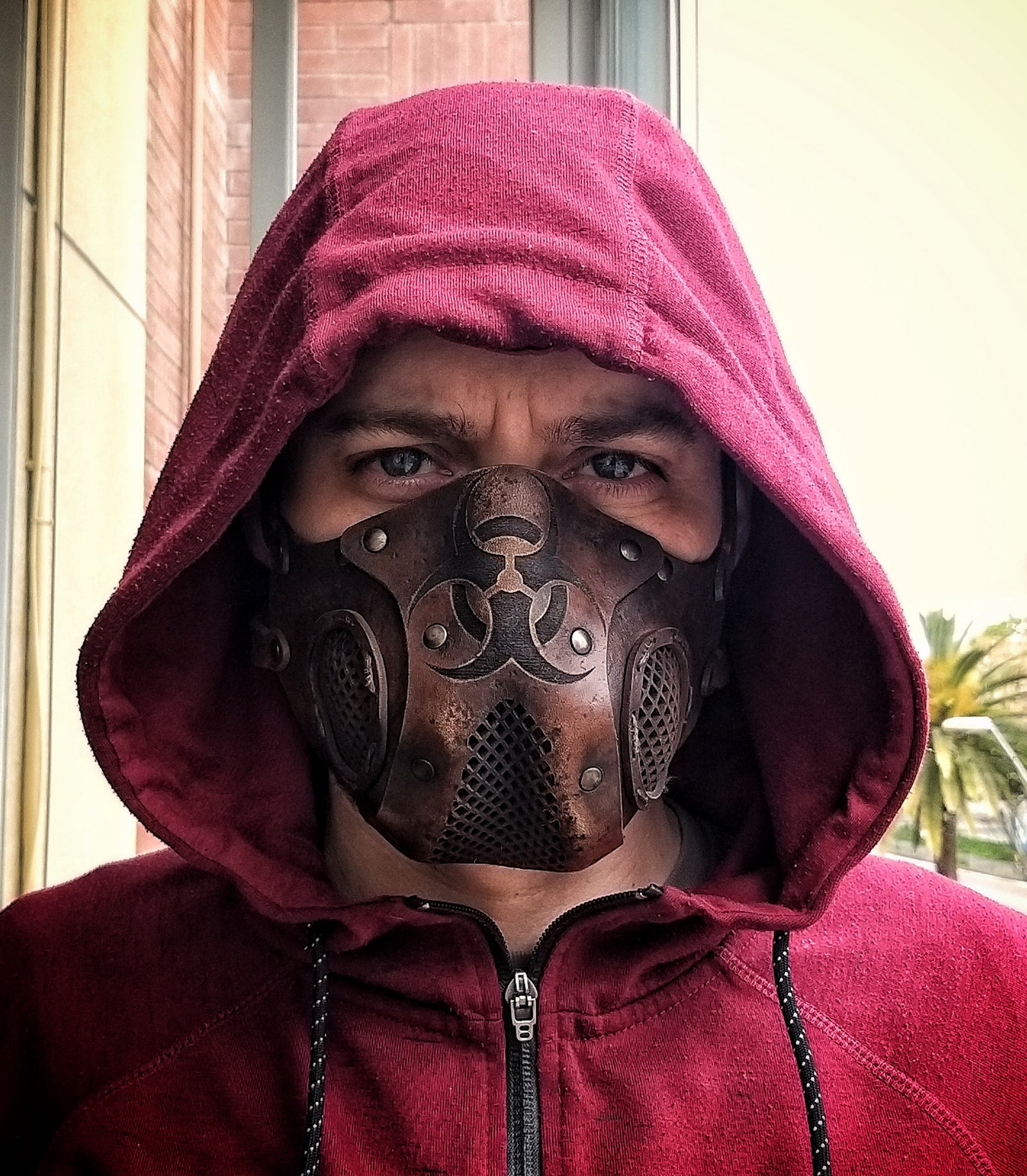 A person wearing a leather face mask with a hood, featuring a biohazard symbol and adjustable straps, in front of a background of foliage. Leather Face Mask with filter pocket Biohazard sign Post Apocalyptic mask for use with disposable face masks Paper towel or filter
