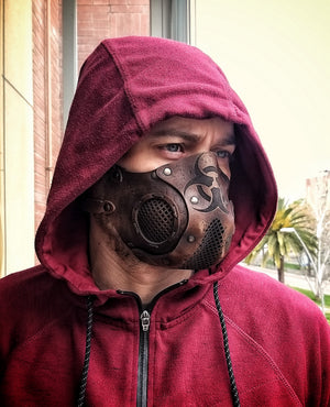 A person wearing a leather face mask with a hood, featuring a biohazard symbol and adjustable straps, in front of a background of foliage. Leather Face Mask with filter pocket Biohazard sign Post Apocalyptic mask for use with disposable face masks Paper towel or filter