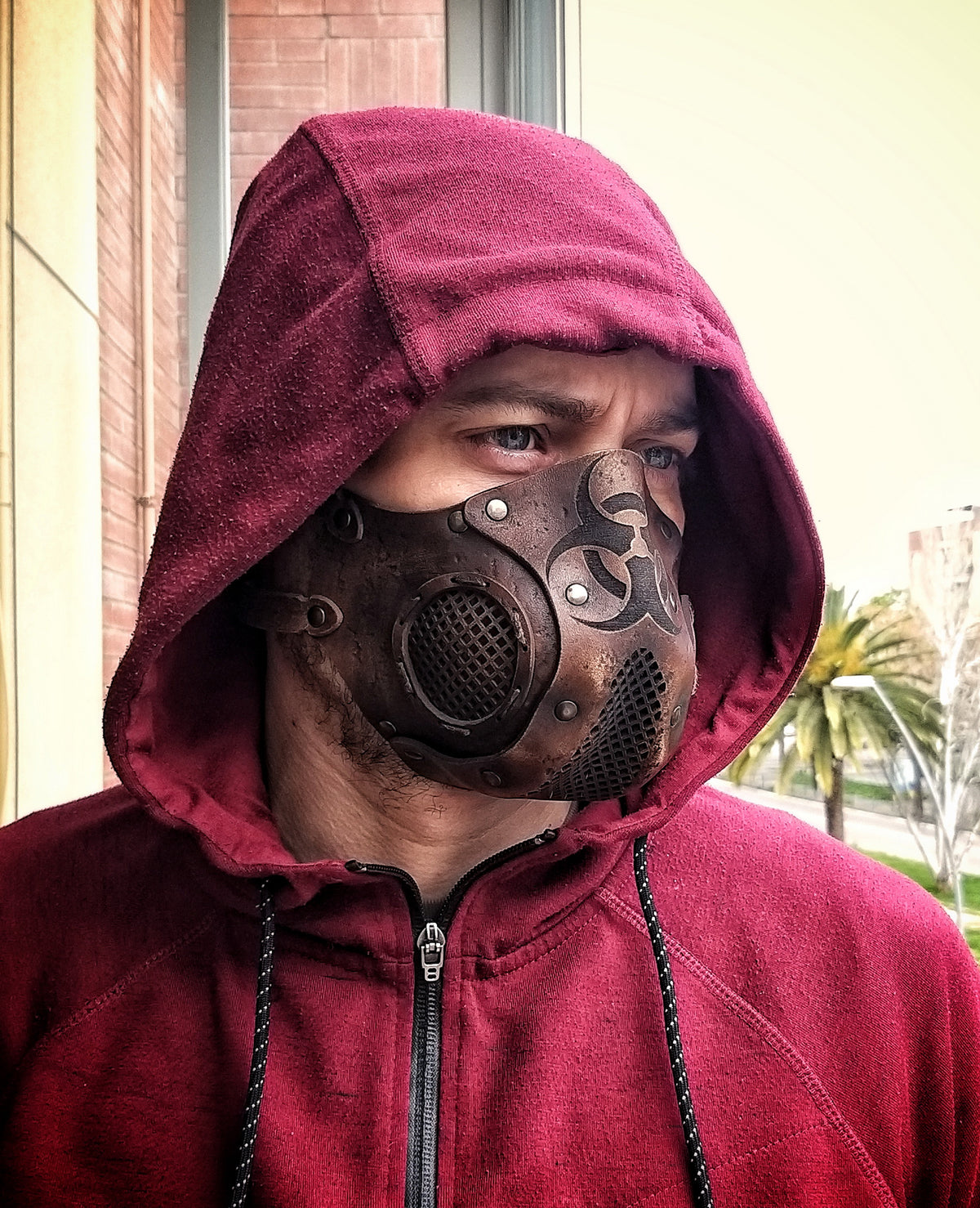 A person wearing a leather face mask with a hood, featuring a biohazard symbol and adjustable straps, in front of a background of foliage. Leather Face Mask with filter pocket Biohazard sign Post Apocalyptic mask for use with disposable face masks Paper towel or filter