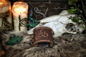 Leather cuff bracelet with the Three Horns of Odin design, placed against a backdrop with candles and decorative foliage. Horns of Odin Leather Cuff Bracelet, Viking Bracer, leather embossed wristband, stamped viking pattern for LARP, cosplay and Fantasy fairs