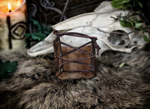 Leather cuff bracelet with the Three Horns of Odin design, placed against a backdrop with candles and decorative foliage. Horns of Odin Leather Cuff Bracelet, Viking Bracer, leather embossed wristband, stamped viking pattern for LARP, cosplay and Fantasy fairs