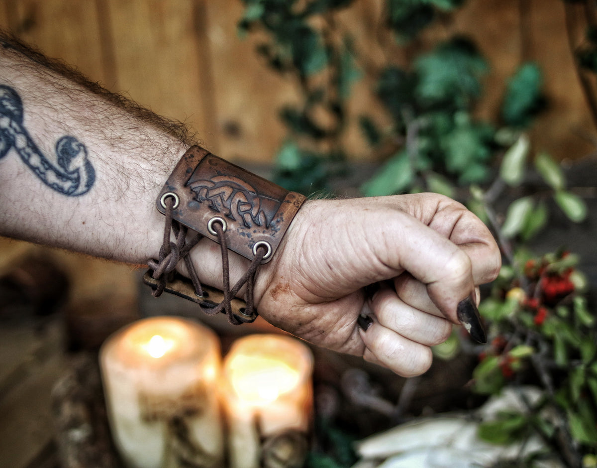 A person's forearm showcasing a leather cuff bracelet with the Three Horns of Odin design, placed against a backdrop with candles and decorative foliage. Horns of Odin Leather Cuff Bracelet, Viking Bracer, leather embossed wristband, stamped viking pattern for LARP, cosplay and Fantasy fairs