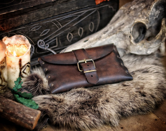 Dark brown fantasy leather pouch resting on fur beside engraved candles and skull. A rustic LARP belt pouch for medieval or Viking reenactment, featuring brass buckle and side rivets. Handmade leather bag for costume or practical use. Scene evokes Nordic or druidic vibe. Alt text designed for SEO and visually descriptive for accessibility.