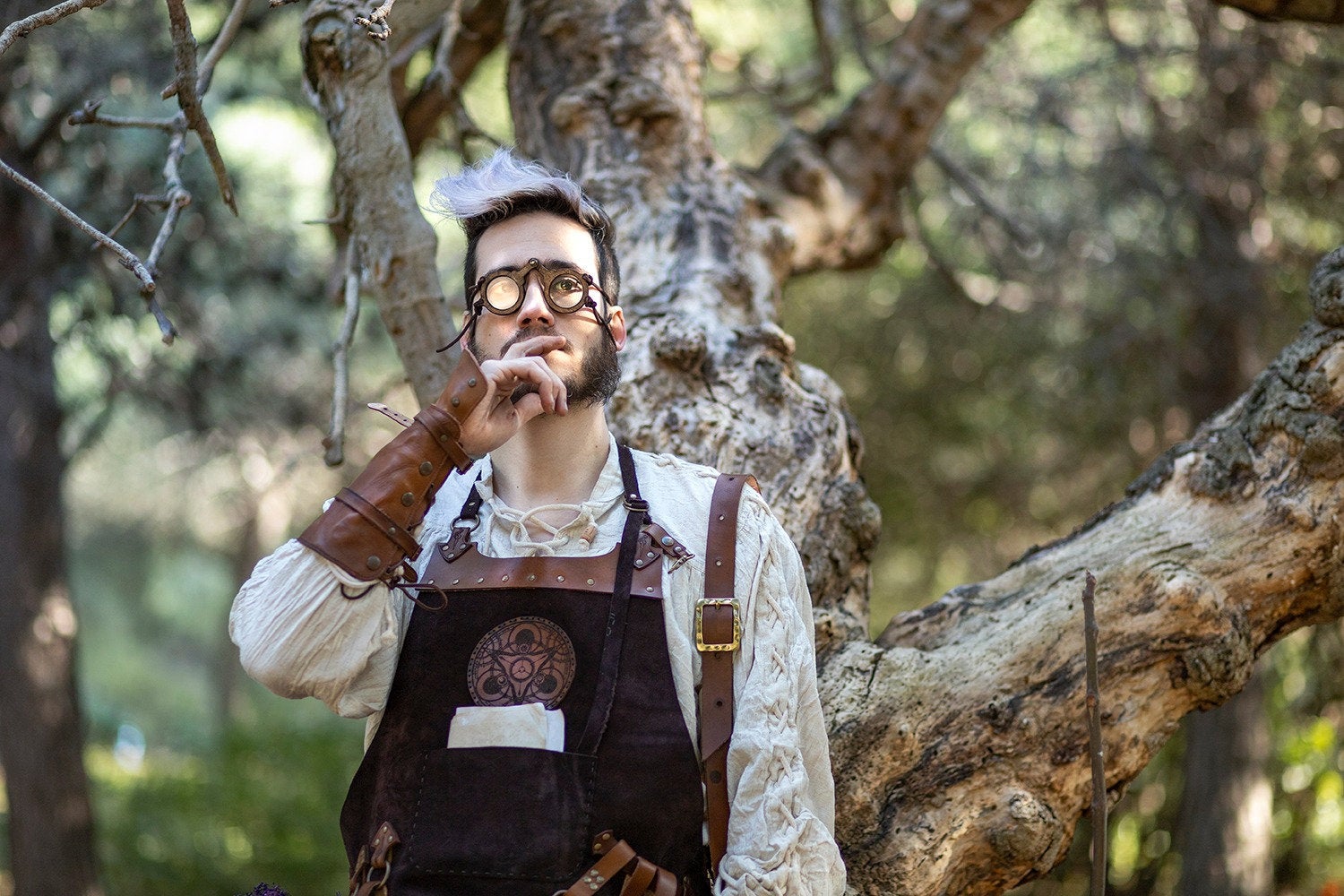 Alchemist character with brown leather gauntlets and handmade medieval spectacles poses thoughtfully under a twisted tree. Perfect for cosplay or LARP wizard costumes, these fantasy eyeglasses add realism and historical flair. Round methacrylate lenses and wooden frame evoke steampunk eyewear style. A person wearing medieval-style wooden eyeglasses with a fantasy character costume, standing in a forest setting.